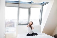 Une jeune femme, assise en tailleur sur son lit avec un casque sur les oreilles, écoute de la musique dans un appartement baigné de lumière