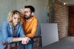 Un couple appuyé sur une barrière dans une maison en construction se montre soucieux face à la situation