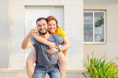 Un jeune homme souriant, qui porte sa compagne à califourchon sur son dos, montre fièrement les clés de leur nouvelle maison