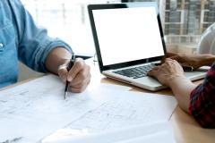 2 personnes travaillent face à face sur un bureau commun, la femme travaille sur son pc, l'homme, sur le plan d'un bâtiment