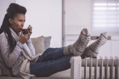 Une femme assise sur son canapé essaie de se réchauffer, en posant ses pieds couverts de grosses chaussettes épaisses contre un radiateur et en buvant une boisson chaude