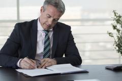Un homme d'âge mûr en costume signe des documents, assis à son bureau