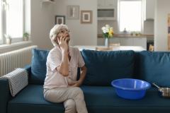 Une femme d'âge mûr, assise sur son canapé, est en communication téléphonique au sujet d'un dégât des eaux dans son salon