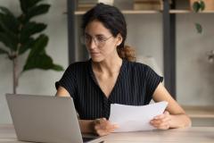 Une femme portant des lunettes et les cheveux attachés consulte des documents sur son pc portable, un document papier dans les mains