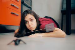 Une jeune femme allongée sur le sol de son salon ouvre de grands yeux face à un gros insecte noir, une tapette à la main