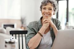 Une femme aux cheveux courts gris, assise devant son pc portable, affiche un sourire satisfait 