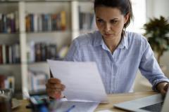 Une femme règle des questions administratives, assise devant son ordinateur portable, un document dans les mains