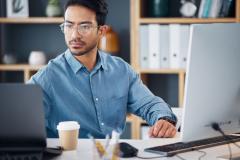 Un homme brun portant des lunettes fait des recherches sur son ordinateur de bureau