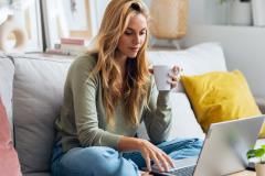 Une jeune femme aux longs cheveux blonds effectue des démarches sur son pc portable, assise en tailleur sur son canapé