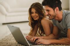Un jeune homme et une jeune femme, à plat ventre sur le tapis de leur salon, font une recherche sur un pc portable