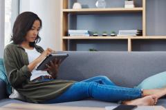 Une jeune femme d'origine asiatique, assise avec les jambes allongées sur son canapé, est concentrée sur le contenu de sa tablette numérique