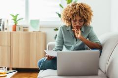 Une jeune femme aux cheveux crépus, assise en tailleur sur son canapé, réalise une étude sur son pc portable