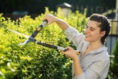 Une jeune femme souriante taille la haie de sa propriété à l'aide d'un taille-haie