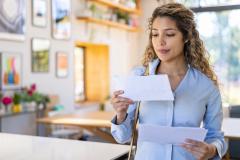 Une jeune femme blonde aux cheveux bouclés lit une lettre qu'elle vient de recevoir, debout dans son salon