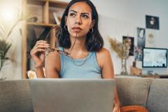Une jeune femme réfléchit dans son salon devant son ordinateur, une paire de lunettes à la main