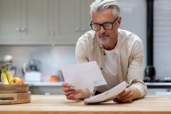 Un homme aux cheveux blancs lit un courrier, en appui sur le plan de travail de sa cuisine