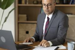 Un homme en costume travaille sur son pc tout en faisant des calculs, assis à son bureau
