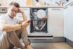 Un homme est assis sur le sol de sa cuisine devant son lave-vaisselle ouvert, son téléphone à la main