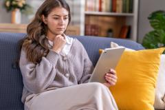Une jeune femme étudie des documents sur une tablette, assise sur son canapé