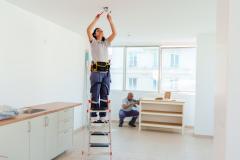 Une électricienne procède à l'installation d'un luminaire dans une cuisine, debout sur un escabeau