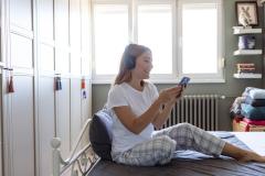 Une jeune femme blonde en pyjama regarde son téléphone, assise sur son lit avec un casque audio sur les oreilles