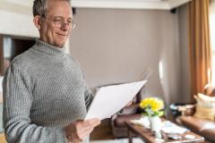 Un homme d'âge mûr examine un document debout dans son salon, avec le sourire