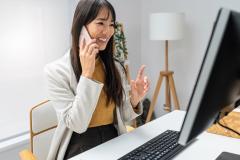 Une femme aux cheveux longs est en pleine conversation téléphonique, assise devant son ordinateur 