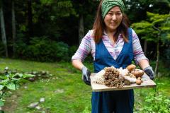Une femme marche dans son jardin, en portant une planche en bois avec les champignons qu'elle vient de ramasser