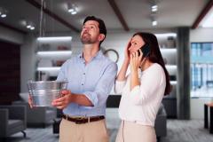 Un homme tient une bassine pour récupérer l'eau qui coule du plafond dans son salon tandis que sa femme à côté de lui téléphone