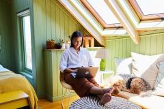 Une jeune femme se détend assise avec son ordinateur sur les genoux dans son studio sous les toits