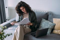Femme travaillant sur son canapé gris, lisant ses documents, avec son ordinateur posé non loin. 
