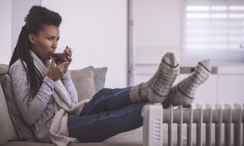 Une femme assise sur son canapé essaie de se réchauffer, en posant ses pieds couverts de grosses chaussettes épaisses contre un radiateur et en buvant une boisson chaude