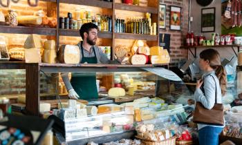 Un homme, debout derrière son comptoir, sert une cliente de sa fromagerie