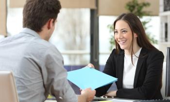 Une jeune femme souriante, assise derrière un bureau, remet un dossier complet à l'homme avec lequel elle s'entretient
