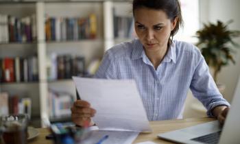 Une femme règle des questions administratives, assise devant son ordinateur portable, un document dans les mains