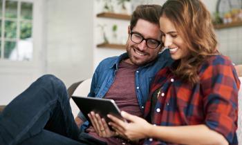 Un couple souriant, installé sur un canapé, réalise une étude sur une tablette