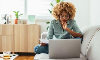 Une jeune femme aux cheveux crépus, assise en tailleur sur son canapé, réalise une étude sur son pc portable
