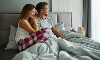 Une femme en pyjama est assise dans son lit contre son compagnon et regarde par la fenêtre en souriant 