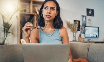 Une jeune femme réfléchit dans son salon devant son ordinateur, une paire de lunettes à la main