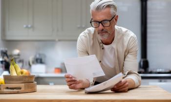 Un homme aux cheveux blancs lit un courrier, en appui sur le plan de travail de sa cuisine