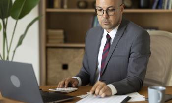 Un homme en costume travaille sur son pc tout en faisant des calculs, assis à son bureau