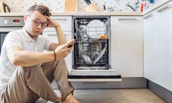 Un homme est assis sur le sol de sa cuisine devant son lave-vaisselle ouvert, son téléphone à la main