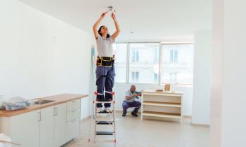 Une électricienne procède à l'installation d'un luminaire dans une cuisine, debout sur un escabeau