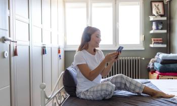 Une jeune femme blonde en pyjama regarde son téléphone, assise sur son lit avec un casque audio sur les oreilles