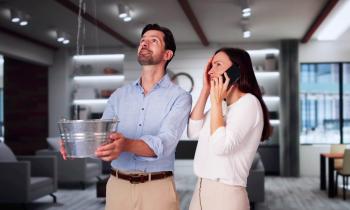 Un homme tient une bassine pour récupérer l'eau qui coule du plafond dans son salon tandis que sa femme à côté de lui téléphone