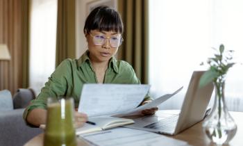 Une femme assise devant son pc portable dans son salon étudie des documents au format papier