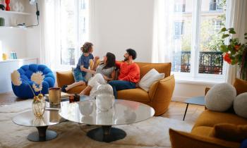Un couple et un enfant rigolent assis sur un canapé jaune moutarde dans un bel appartement parisien. 