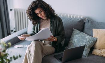 Femme travaillant sur son canapé gris, lisant ses documents, avec son ordinateur posé non loin. 