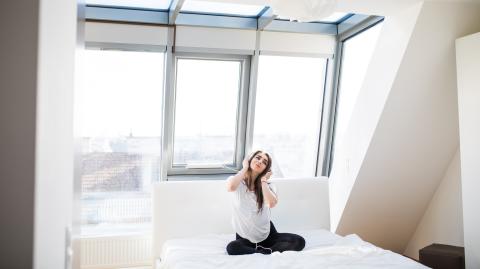 Une jeune femme, assise en tailleur sur son lit avec un casque sur les oreilles, écoute de la musique dans un appartement baigné de lumière