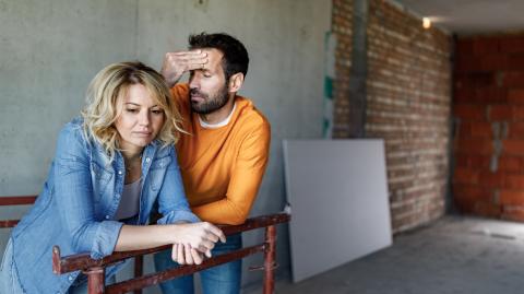 Un couple appuyé sur une barrière dans une maison en construction se montre soucieux face à la situation